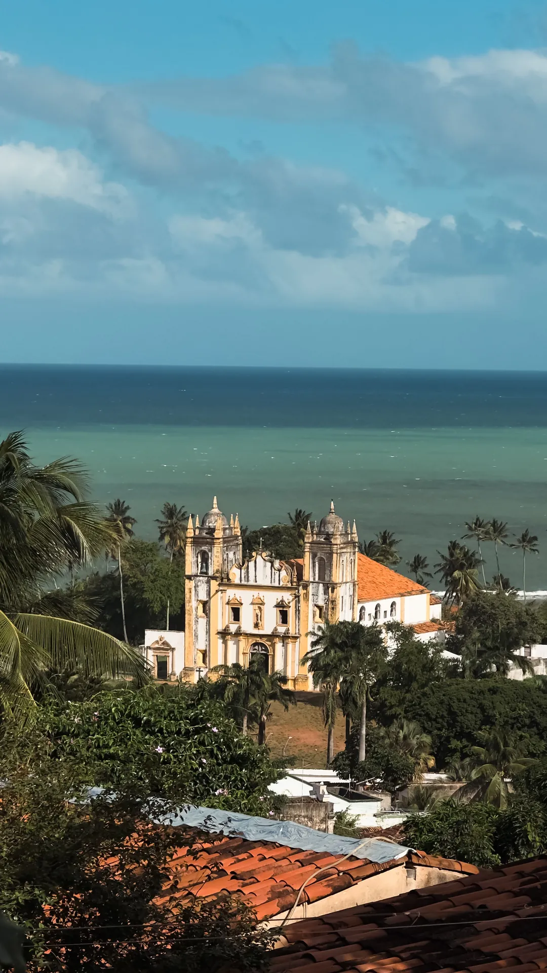 Igreja histórica de Olinda entre palmeiras, com o mar verde e o céu do fim de tarde ao fundo