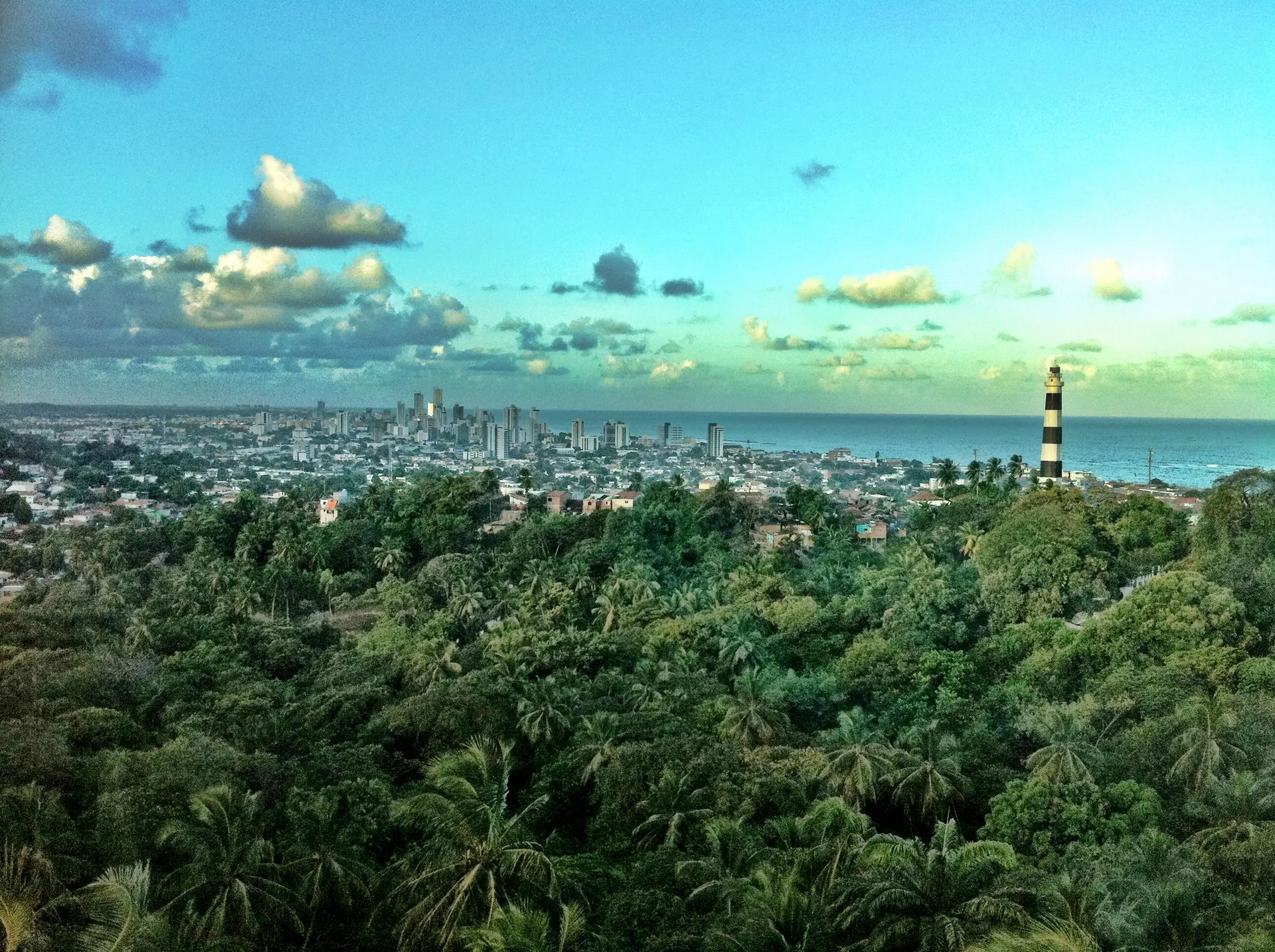 Vista do alto de Olinda com o farol, o casario entre coqueiros e a cidade do Recife no horizonte