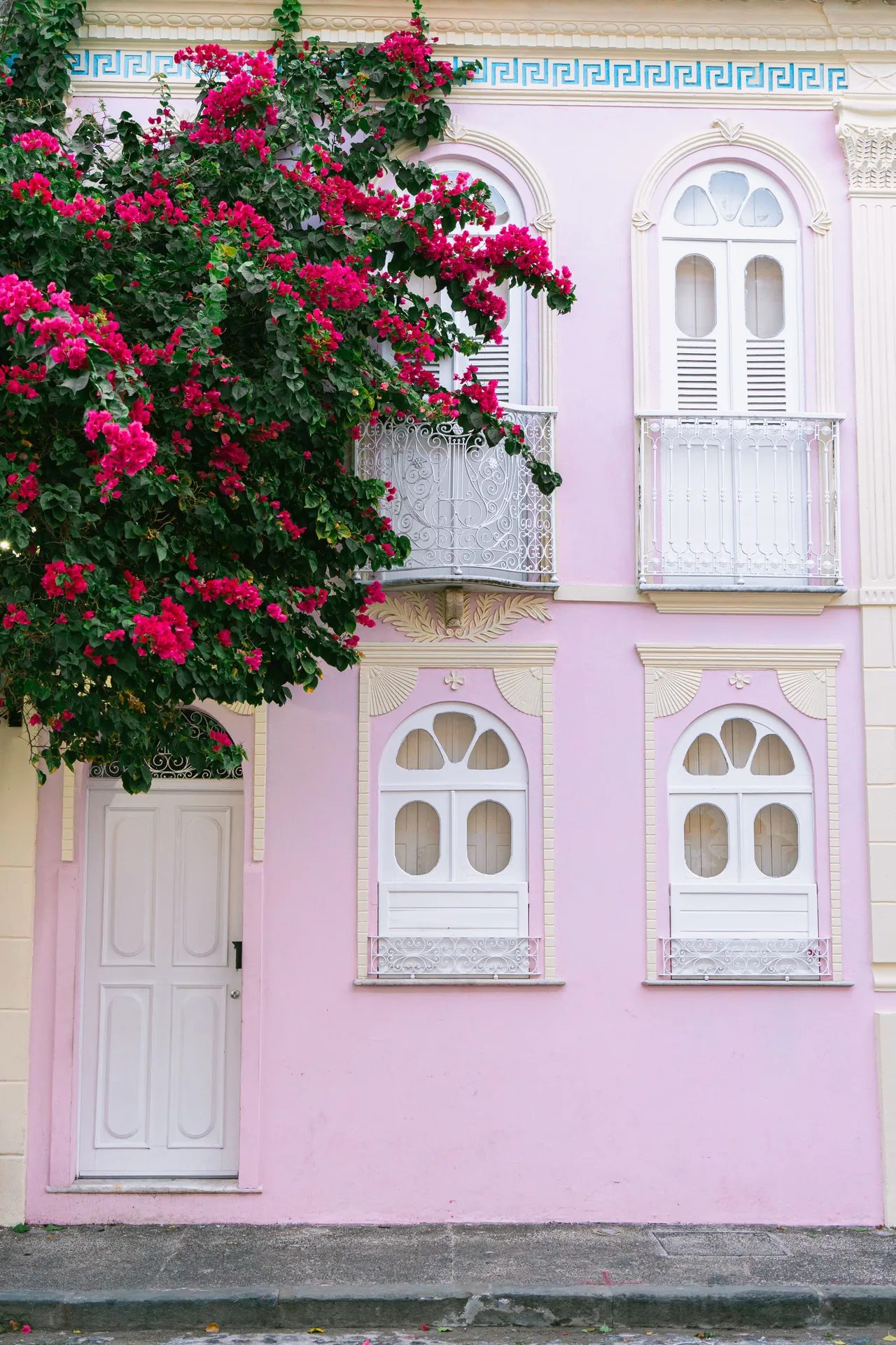 Fachada de casa rosa com janelas brancas em arco e uma buganvília carregada de flores rosa sobre a sacada