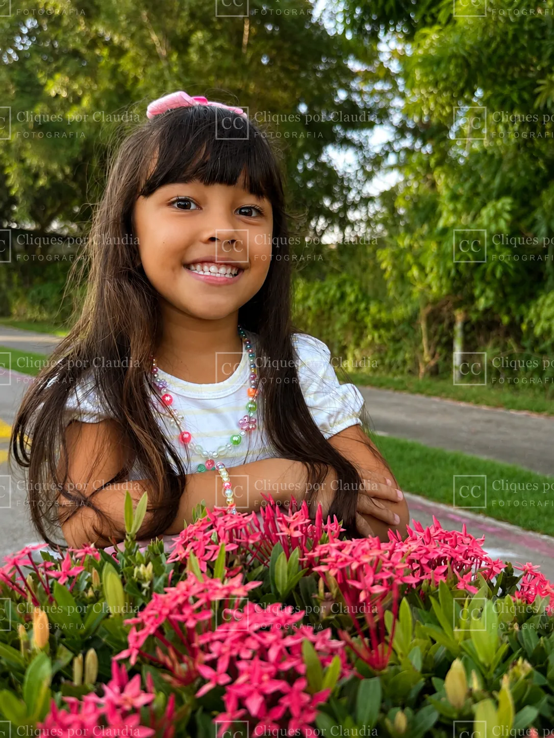 Maria Lívia sorrindo apoiada num canteiro de flores vermelhas na Praia do Paiva
